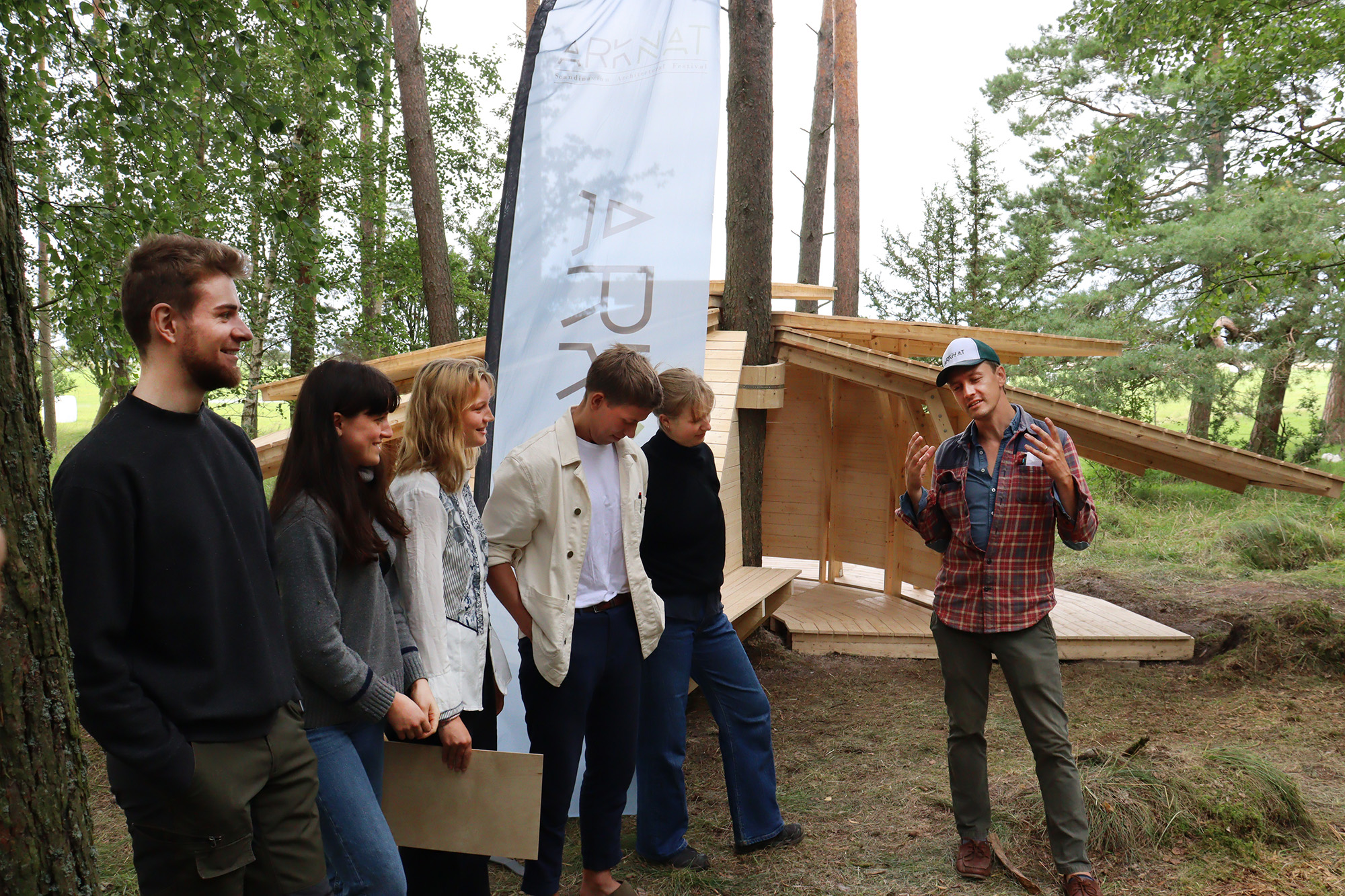 Fem studenter lyssnar på projektledaren Martin Björklund. Bakom dem syns vindskyddet i trä